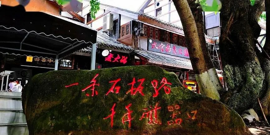 A mossy boulder at Ciqikou Ancient Town carved in red with the line 'One flagstone road, a thousand years of Ciqikou', beside shopfronts and a large tree